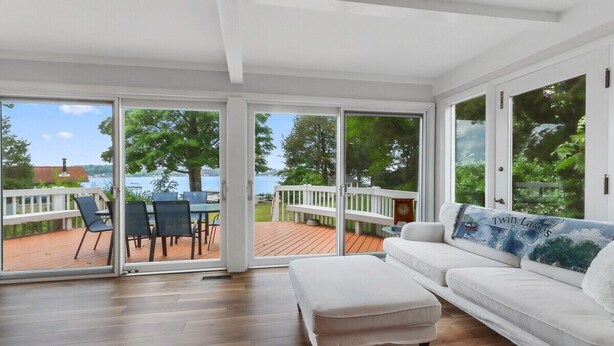 A spacious room with a white couch, a wooden floor, and large sliding glass doors leading to an outdoor deck with a view of a lake and trees.