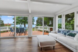 Une pièce spacieuse avec un canapé blanc, un plancher en bois et de grandes portes-fenêtres coulissantes donnant sur une terrasse extérieure d’où l’on peut admirer un lac et des arbres.