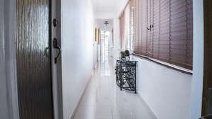Un couloir avec une porte à gauche, une fenêtre avec des stores à droite et une petite table avec une plante au centre.