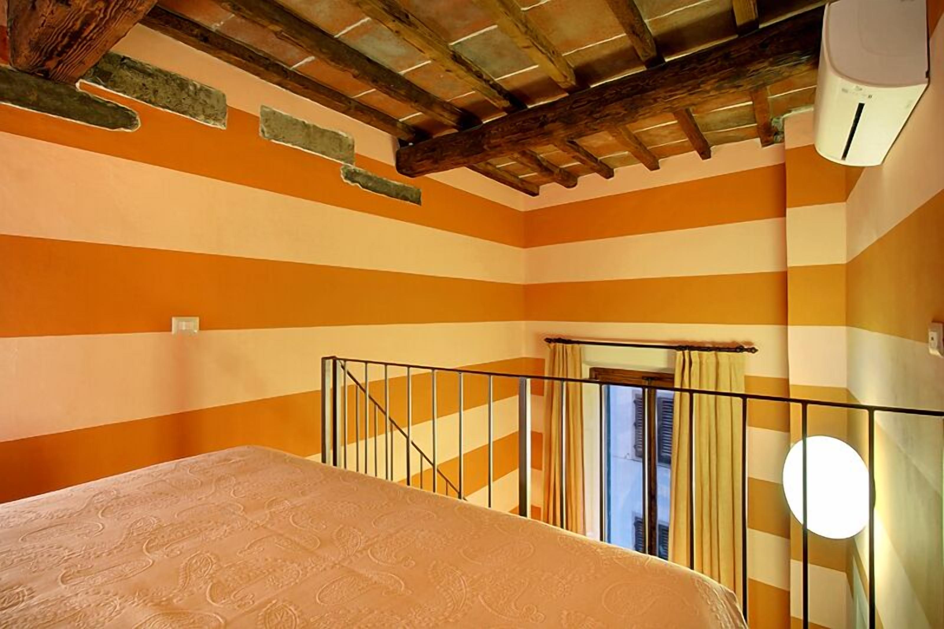 Una habitación con paredes a rayas anaranjadas y blancas, techo de madera, cama con cubierta estampada y ventana con cortinas.