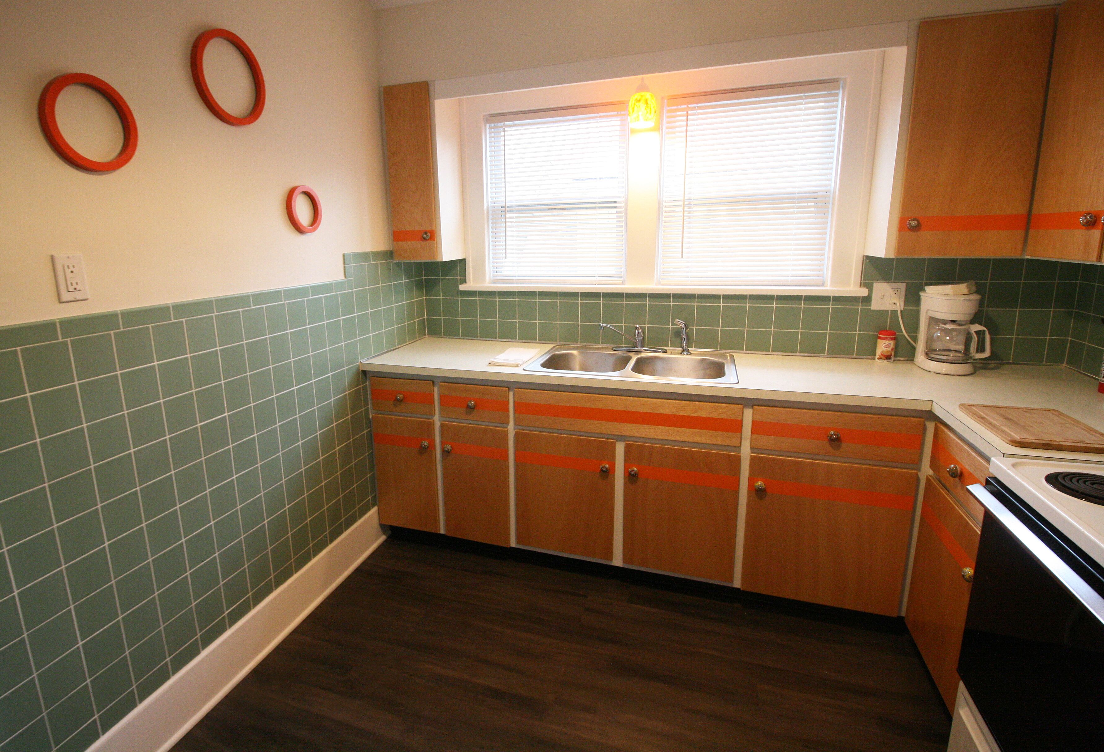 A kitchen with green tiled walls, wooden cabinets, a double sink, and a window with blinds.