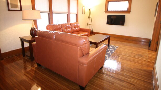 A living room with a leather sofa, a small table, a floor lamp, and a TV mounted on the wall. - Safe, Clean & Spacious, Great for Families (Niagara Falls)