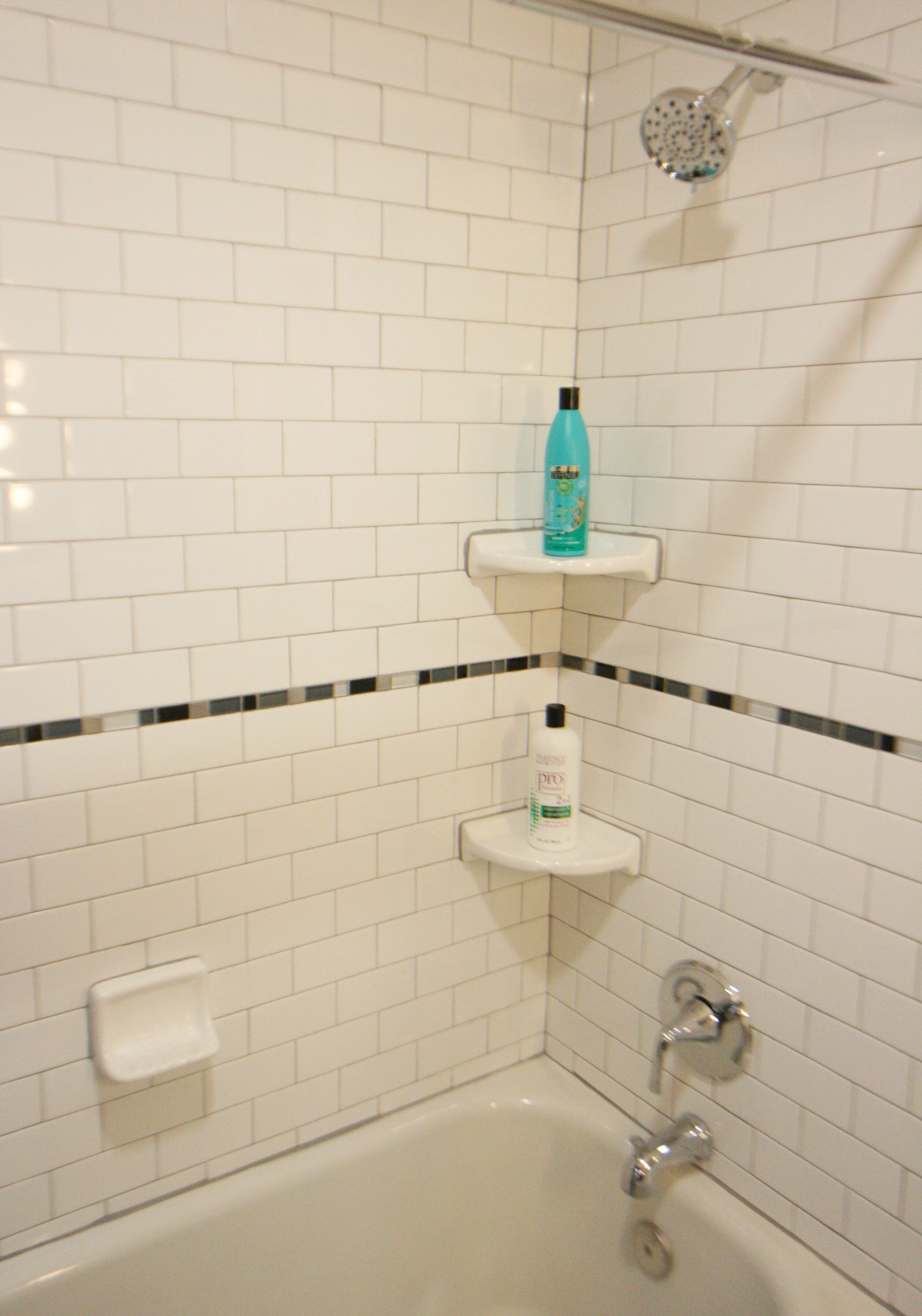 A bathroom with white tiled walls, a bathtub, a showerhead, and two shelves with toiletries.