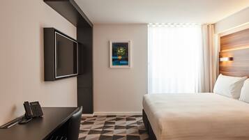 A hotel room with a bed, a desk with a telephone, a flat-screen TV mounted on the wall, and a framed picture on the wall.