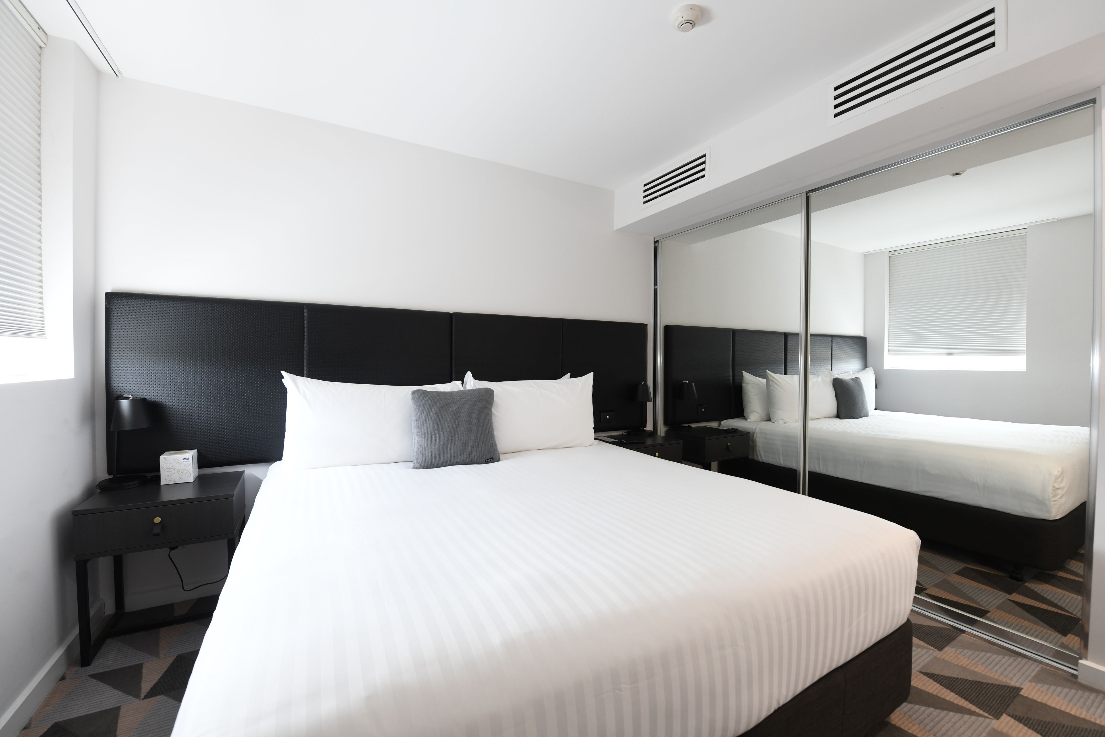 A modern hotel room with a large bed, a bedside table, a nightstand, a mirror, and a window with blinds.