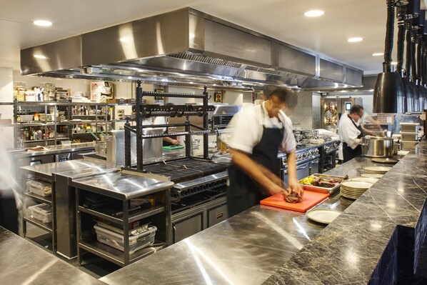 A commercial kitchen with stainless steel appliances, a chef preparing food, and a long counter with plates and utensils. - The Gem Hotel (Griffith)