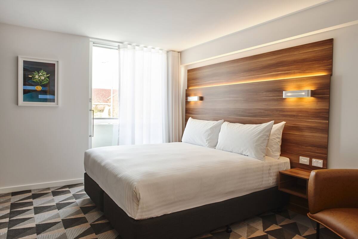 Una habitación de hotel con una cama grande, cabecera de madera, mesita de noche, una silla y una ventana con cortinas.