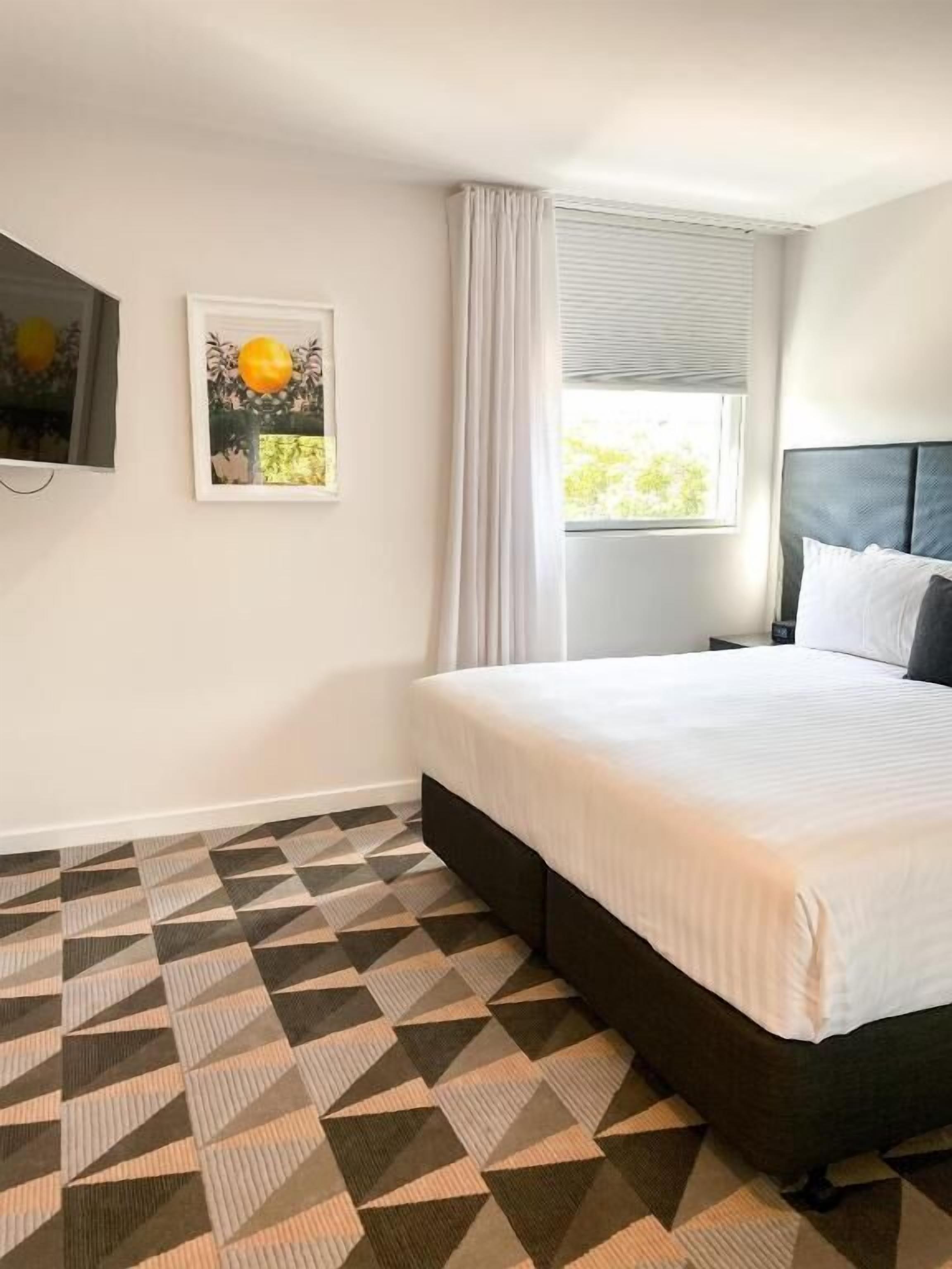 A bedroom with a bed, a window with curtains, a wall-mounted TV, and a geometric patterned floor.