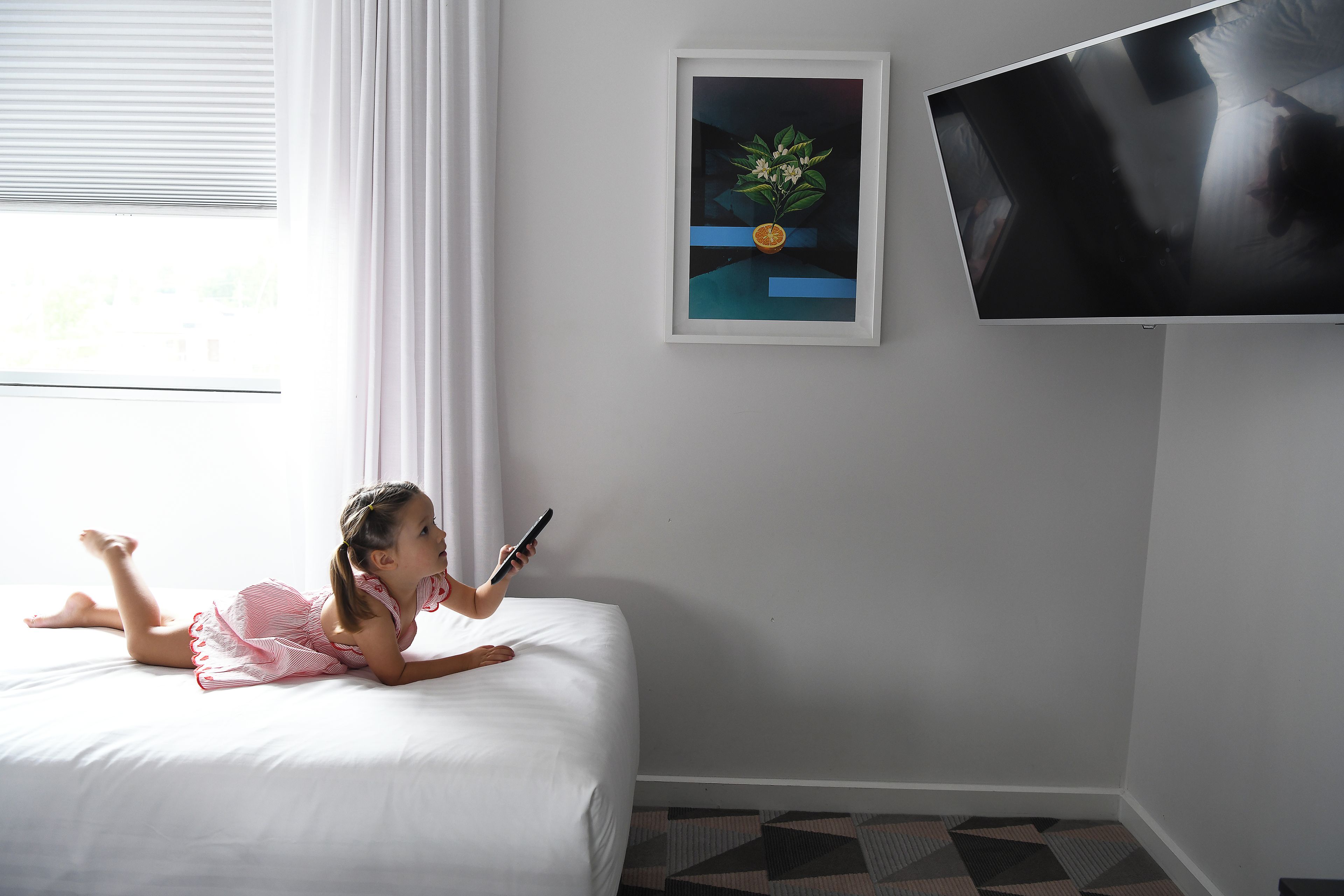 A child lying on a bed with a smartphone, a framed picture on the wall, and a television mounted on the wall.