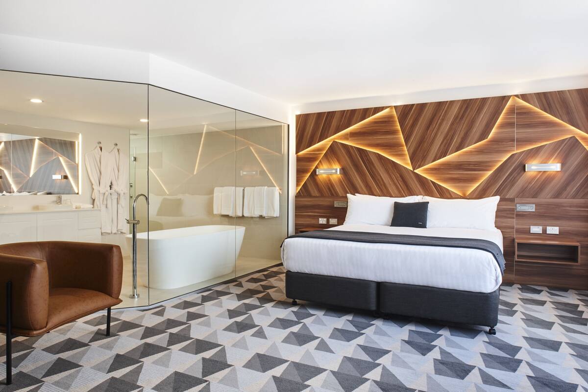 Habitación de hotel moderna con una cama grande, cabina de ducha de vidrio, un sillón de cuero marrón y piso con un diseño geométrico.