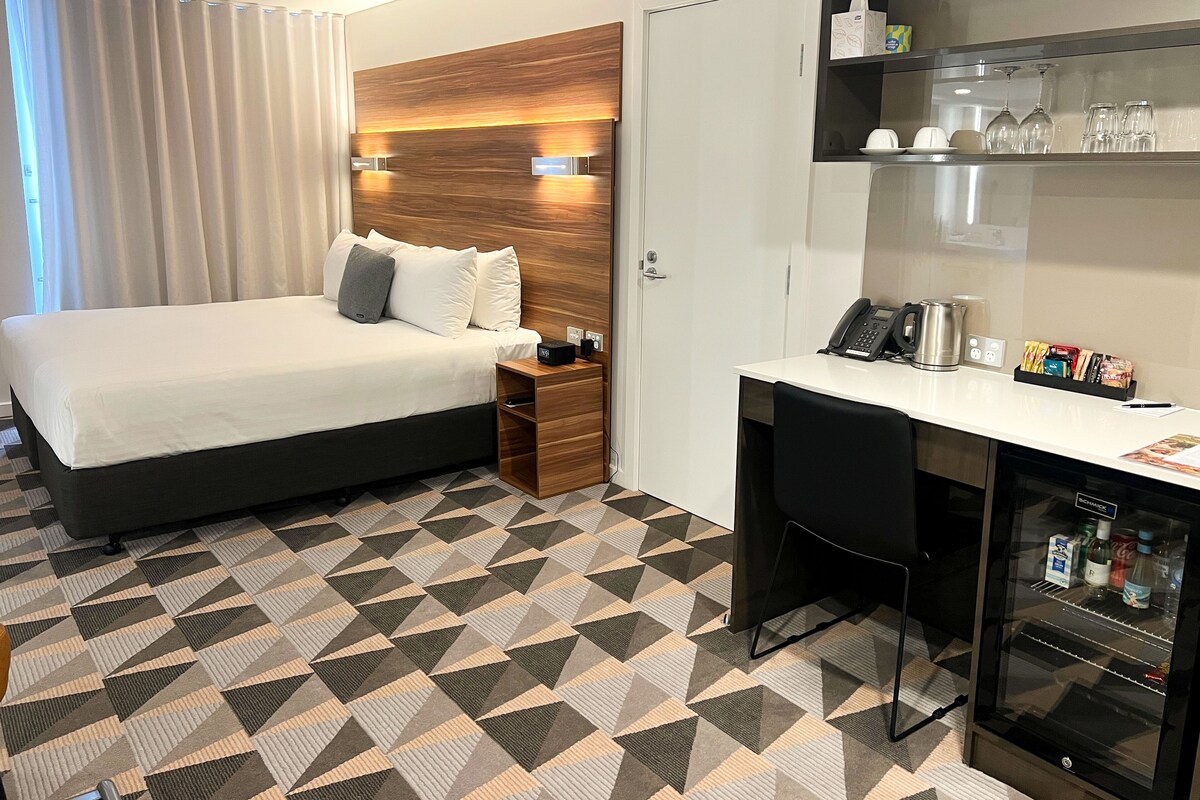 A hotel room with a bed, a desk, a minibar, and a coffee machine.