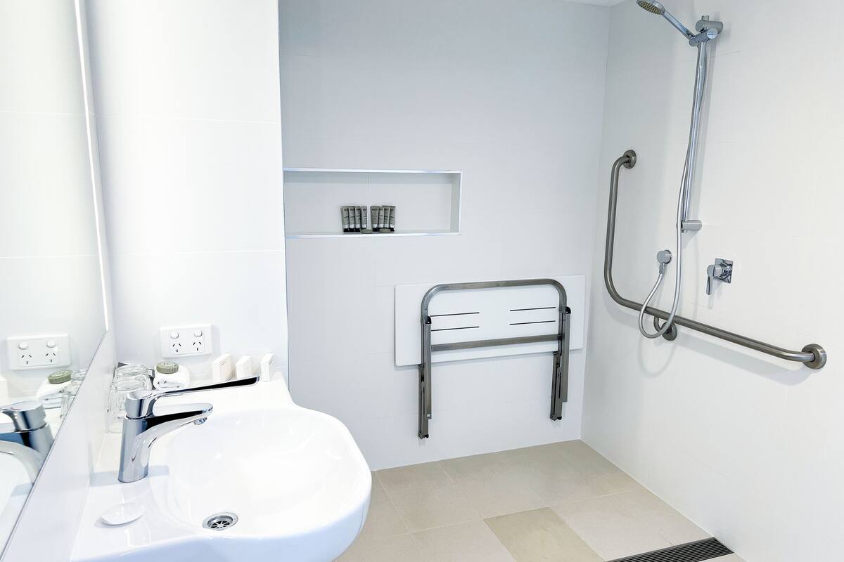 Un baño moderno con un lavabo blanco, una ducha con barra de apoyo y un estante con artículos de aseo.