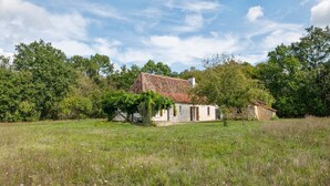 A rustic house with a tiled roof, surrounded by a grassy field and dense trees.