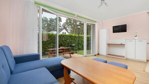 A modern living room with a wooden table and blue sofas, a TV mounted on the wall, and a sliding glass door leading to an outdoor patio with a barbecue grill. - Large, comfortable holiday homes close to the sea (Niechorze)