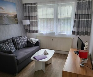 Un salon moderne avec un canapé gris, une table basse blanche, un bureau en bois avec une lampe rouge et un radiateur.