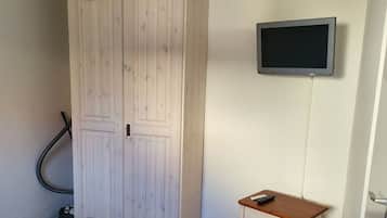 Una habitación con un armario blanco grande, un escritorio de madera pequeño con un televisor montado en la pared y una aspiradora en la esquina.