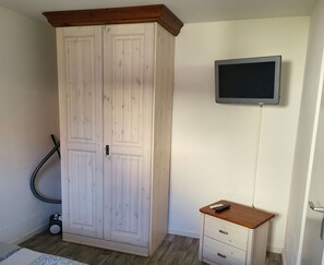 A room with a large white wardrobe, a small wooden desk with a TV mounted on the wall, and a vacuum cleaner in the corner.