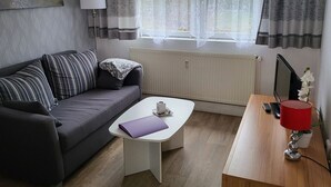 Uma sala de estar moderna com um sofá cinzento, uma mesa de centro branca, uma escrivaninha de madeira com uma luminária vermelha e um radiador.