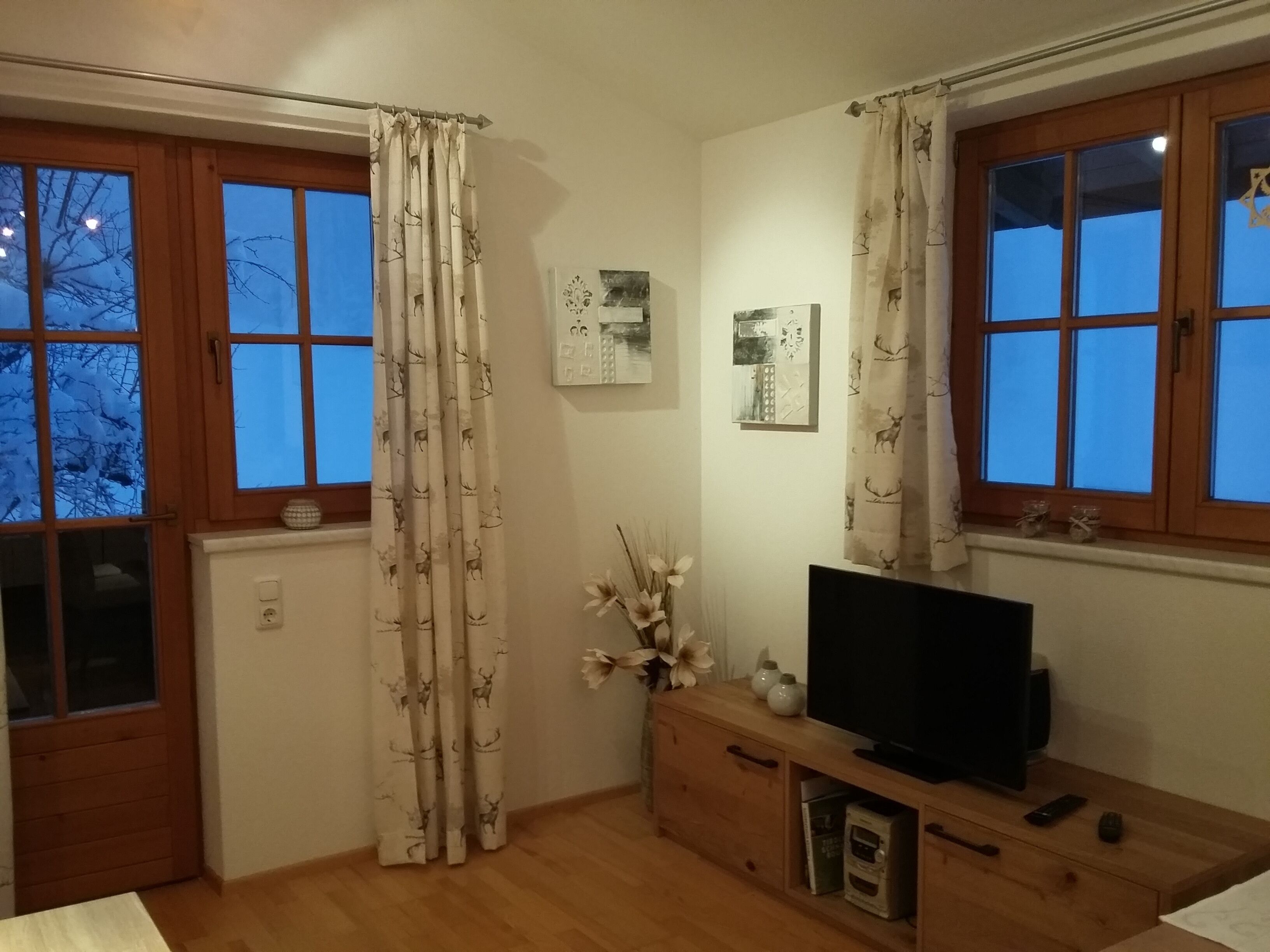 Una habitación con pisos de madera, un televisor sobre un mueble de madera, una ventana con cortina y un cuadro colgado en la pared.