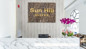 Un comptoir d’accueil avec un plateau en marbre, une enseigne fixée au mur portant l’inscription « Sun Hill Hotel », et une plante en pot ornée de fleurs violettes.