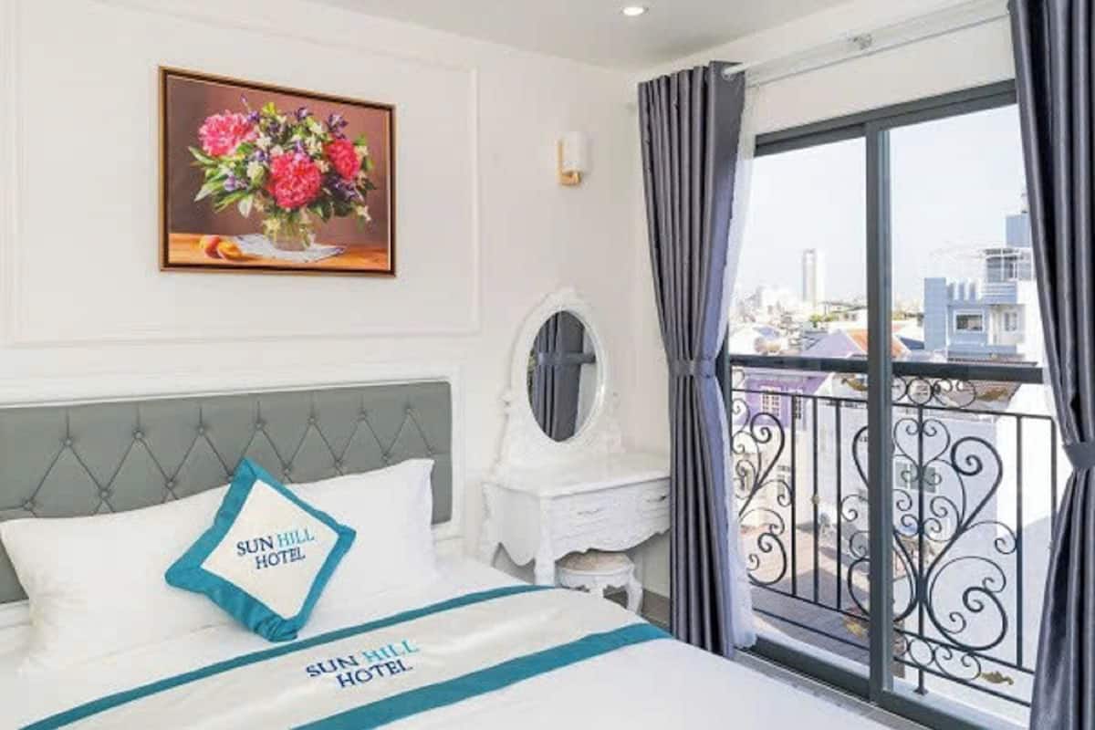 Ein Hotelzimmer mit einem Bett, einem Nachttisch, einem Spiegel, einem gerahmten Blumenbild und einem Balkon mit Blick auf die Stadt.