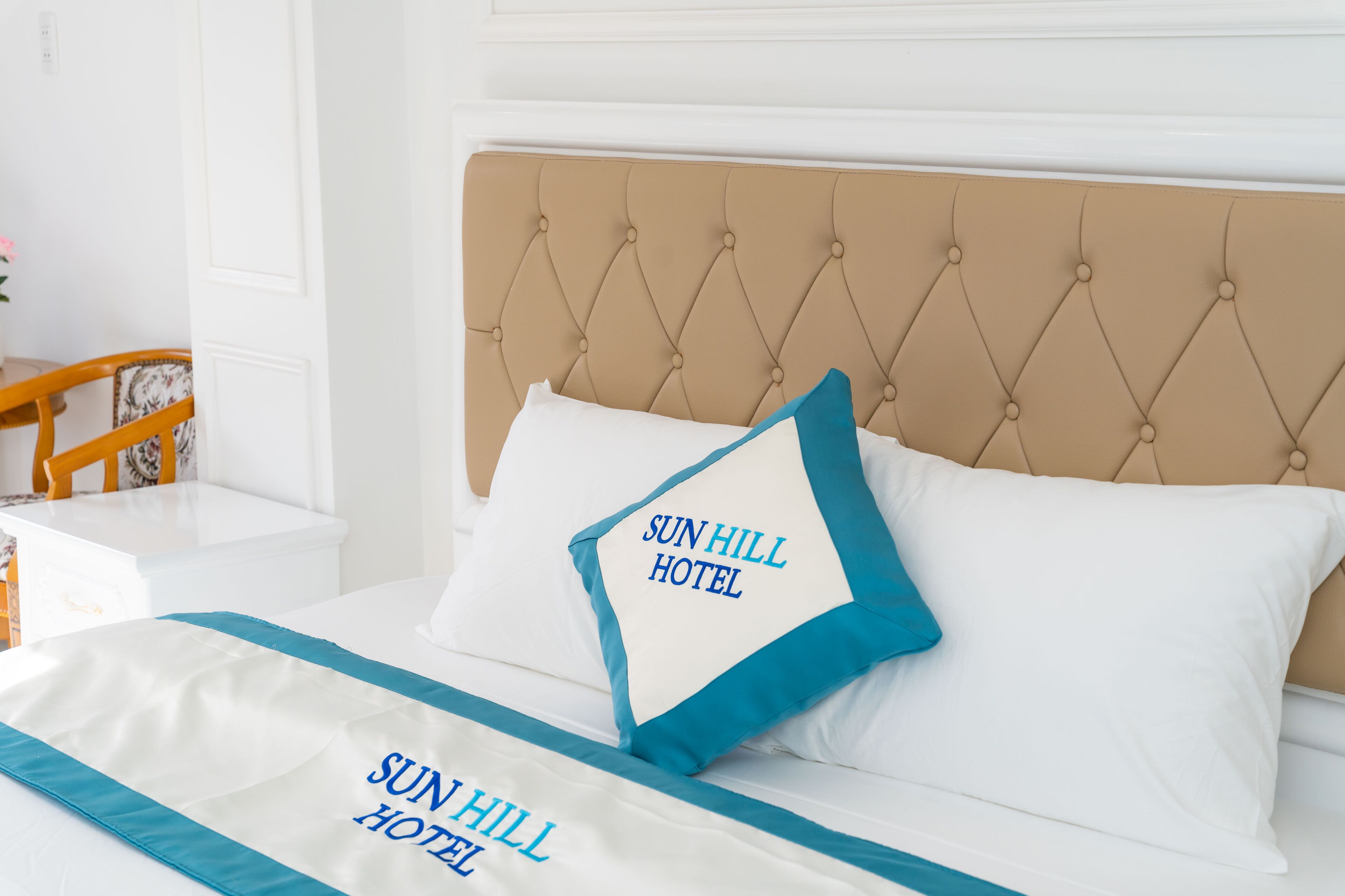 A hotel room with a tufted headboard, a bed with white and blue bedding, and a pillow with "Sun Hill Hotel" text.