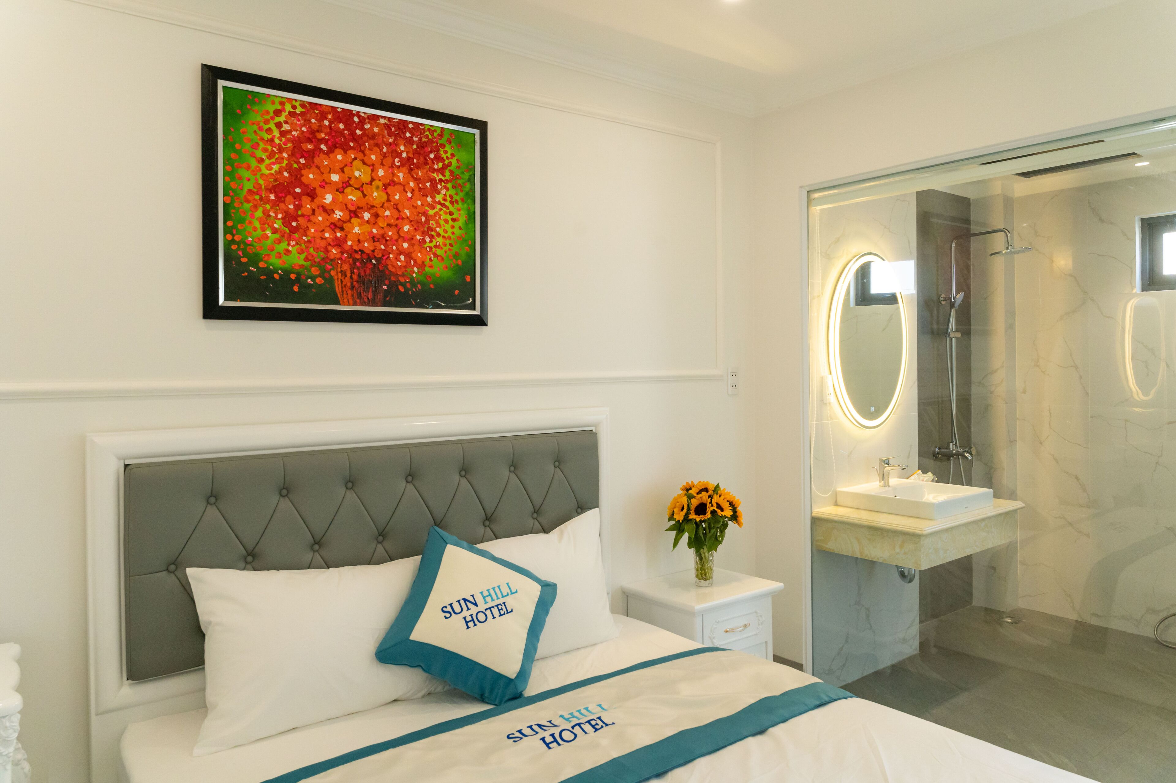 Een hotelkamer met een bed, een nachtkastje met een bloevas, een badkamer met spiegel en wastafel, en een lijst met kunstwerk aan de muur.