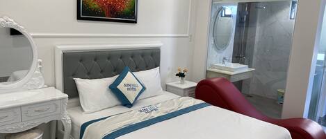 Habitación de hotel con cama, mesita de noche, espejo, una silla roja, baño con cabina de ducha de vidrio y un cuadro en la pared.