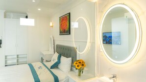 Une chambre d’hôtel avec un lit, une table de chevet ornée d’un vase de tournesols, un miroir rond avec un éclairage intégré, une armoire blanche et un téléviseur fixé au mur.