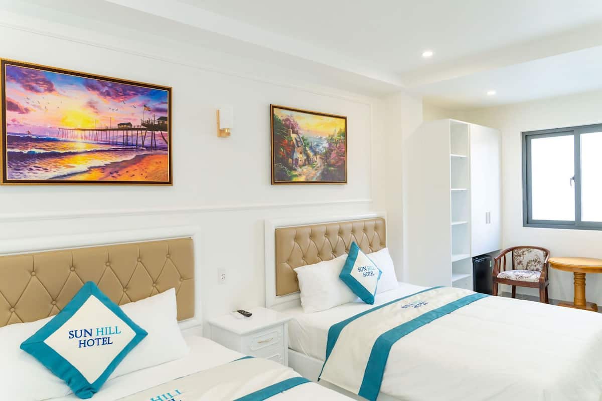 Ein Hotelzimmer mit zwei Betten, einem Schreibtisch und einem Stuhl. An den Wänden befinden sich zwei Bilder, ein weißer Kleiderschrank und ein Fenster.