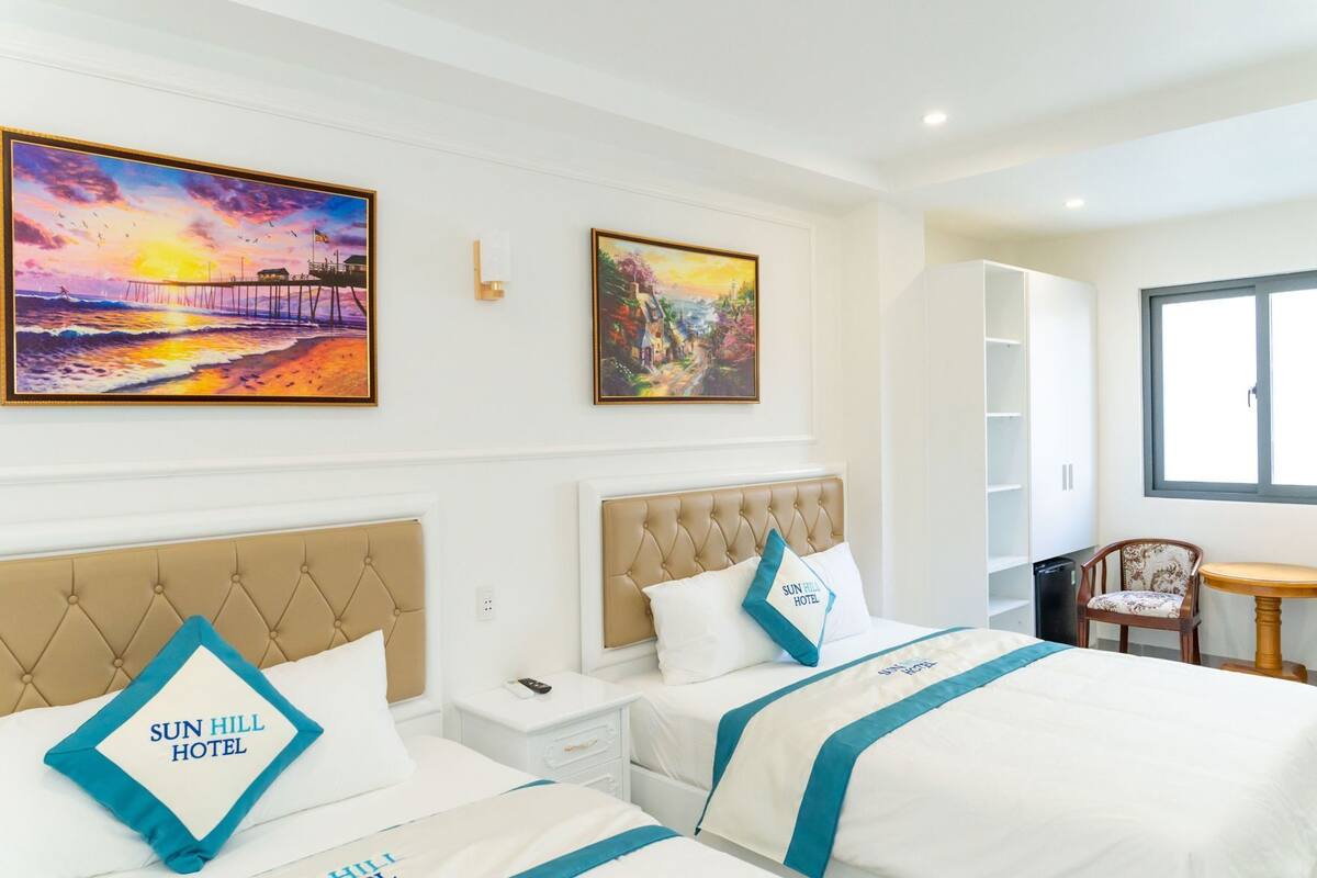Habitación de hotel con dos camas, un escritorio y una silla. Hay dos cuadros en la pared, un armario blanco y una ventana.