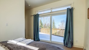 Une chambre dotée d’un grand lit, d’une porte-fenêtre coulissante donnant sur un balcon, et offrant une vue sur les arbres et une terrasse en bois.