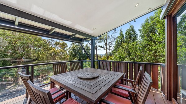 Una mesa de madera con cojines rojos en un balcón con barandas de vidrio, con vistas a una zona boscosa.