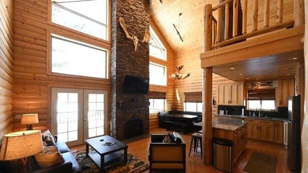 Interior espacioso de una cabaña de troncos, con chimenea, pisos de madera, zona de comedor y cocina.