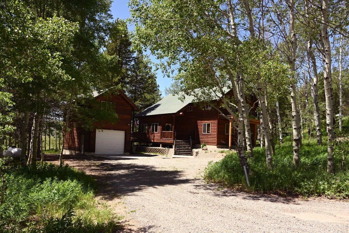A two-story log cabin with a garage, surrounded by trees and a gravel driveway.