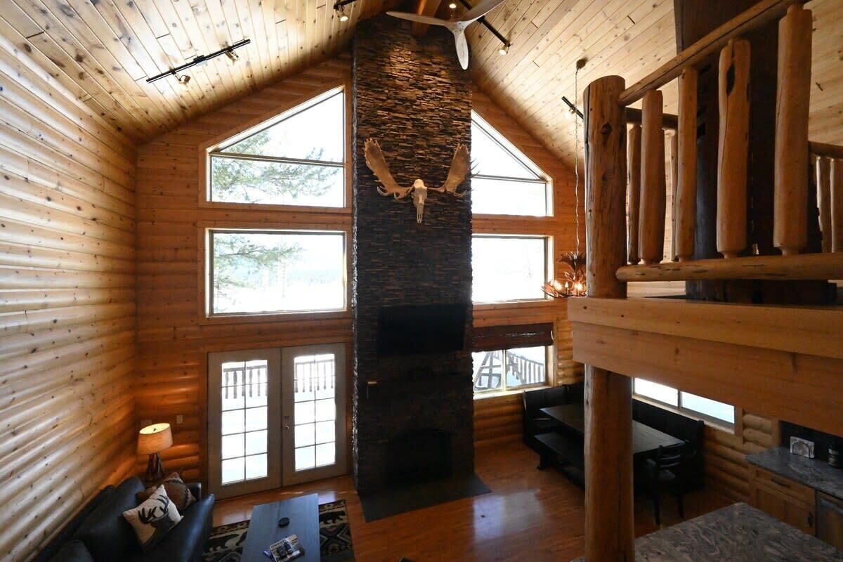 A log cabin interior with a stone fireplace, wooden walls, large windows, and a loft area.
