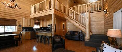 A log cabin interior with a dining area, kitchen, and a staircase leading to an upper level.