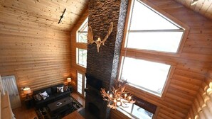 Interior de una cabaña de troncos con chimenea de piedra, paredes de madera y ventanas amplias.