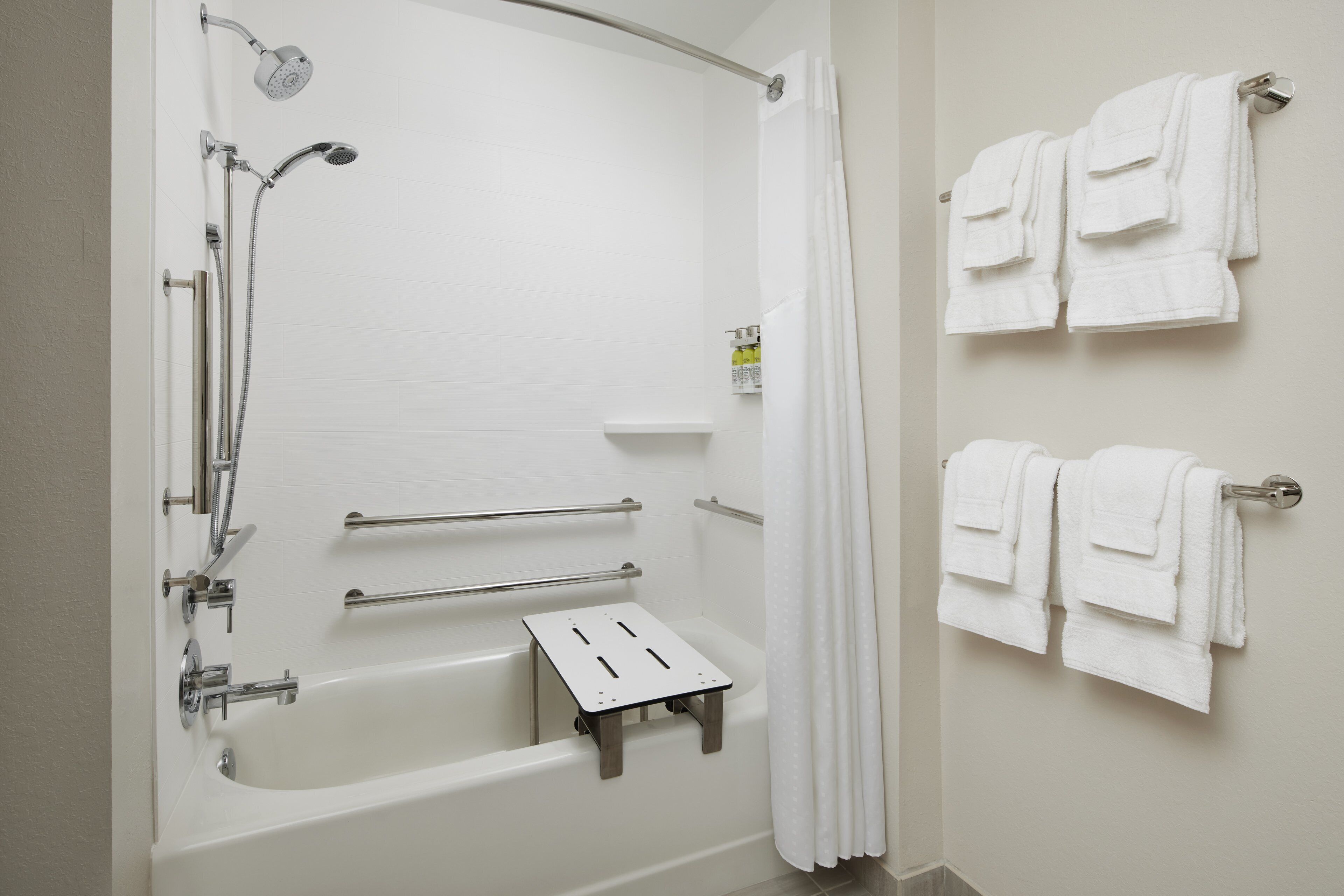 A modern bathroom with a bathtub, shower, and handrails. There are towels on a rack and a shower curtain.