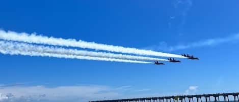 A clear blue sky with a jet formation flying overhead and a pier extending into the water.