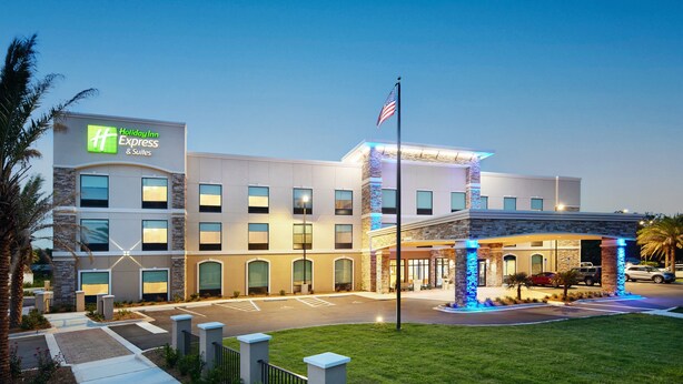 A Holiday Inn Express & Suites building with a parking lot, a flag, and a well-lit entrance. - HOLIDAY INN EXPRESS & SUITES GULF BREEZE - PENSACOLA AREA by IHG (Gulf Breeze)