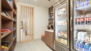 A clean, well-organized snack and beverage area with shelves stocked with various snacks and drinks, and a refrigerator with beverages. - HOLIDAY INN EXPRESS & SUITES GULF BREEZE - PENSACOLA AREA by IHG (Gulf Breeze)