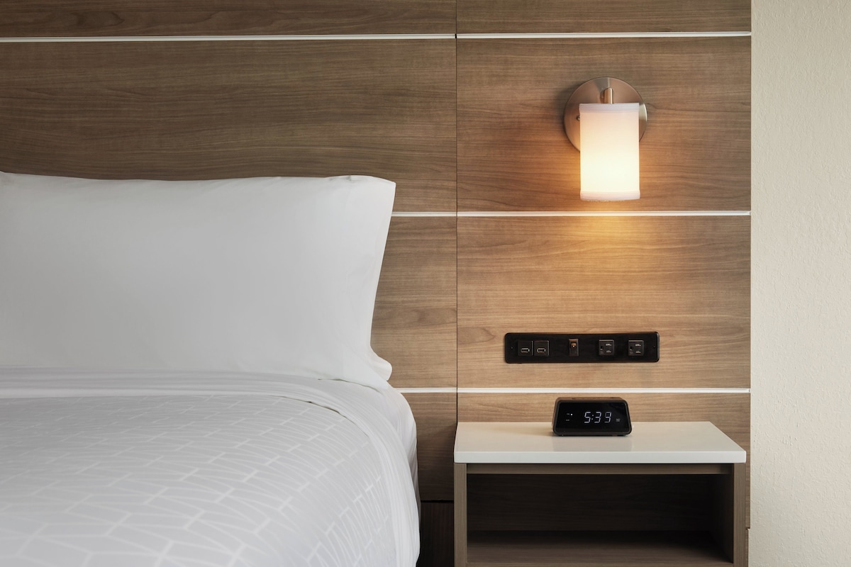 Un lit bien fait, avec une couette blanche et des oreillers, une table de chevet dotée d’une horloge numérique et un luminaire fixé au mur.