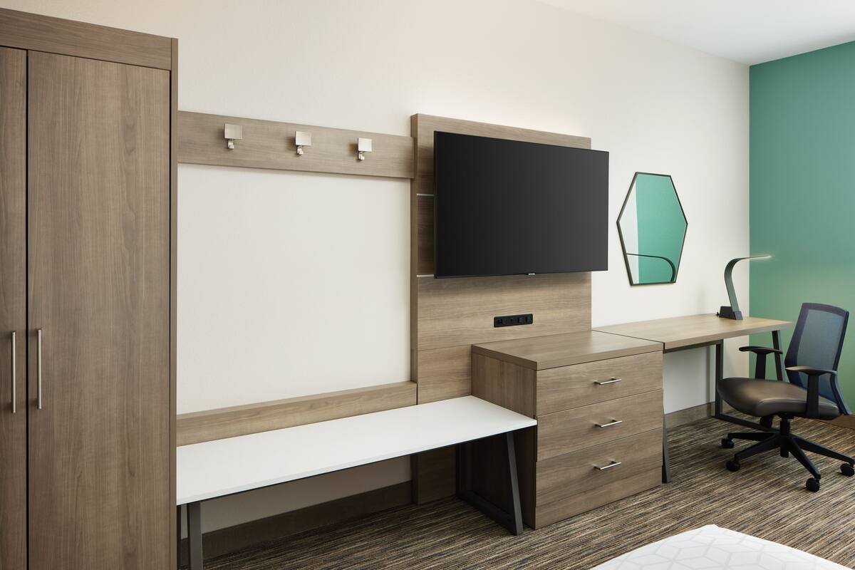 Une chambre d’hôtel moderne avec une armoire en bois, un bureau doté d’un téléviseur à écran plat, un banc, une chaise et un mur d’accent vert.