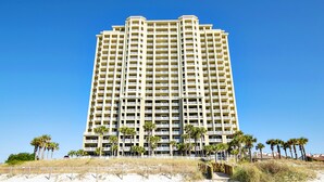 A tall, multi-story hotel with balconies, located on a beach with palm trees and sandy ground. - 🌞Beach Front-Corner Unit-Grand Panama 1-409🌞 (Panama City Beach)