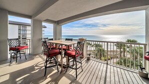 A balcony with a wooden table and chairs, overlooking a beach with palm trees and a clear sky. - 🌞Beach Front-Corner Unit-Grand Panama 1-409🌞 (Panama City Beach)