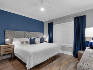 Une chambre à coucher comprenant un lit, des tables de chevet, une lampe, une fenêtre avec des rideaux et un mur orné d’une tête de lit à motifs.