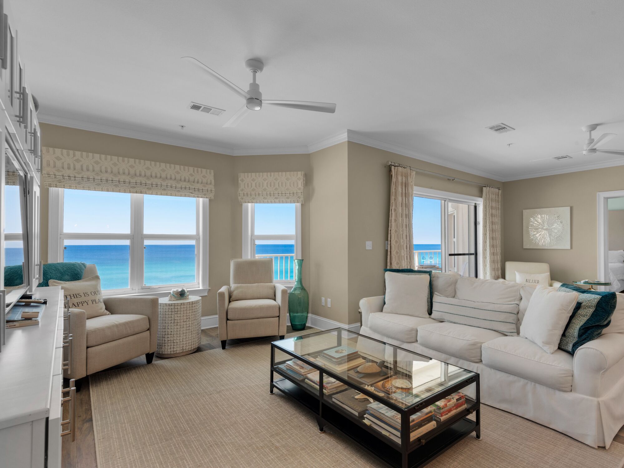 A spacious living room with a large window offering a view of the ocean, a ceiling fan, and a glass coffee table with books.