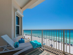 A balcony with a view of a beach, a lounge chair, and a small table with a towel and a bottle. - Gulf Front! Designer Decorated! Perfect View! Community Pool and Hot Tub (Destin)