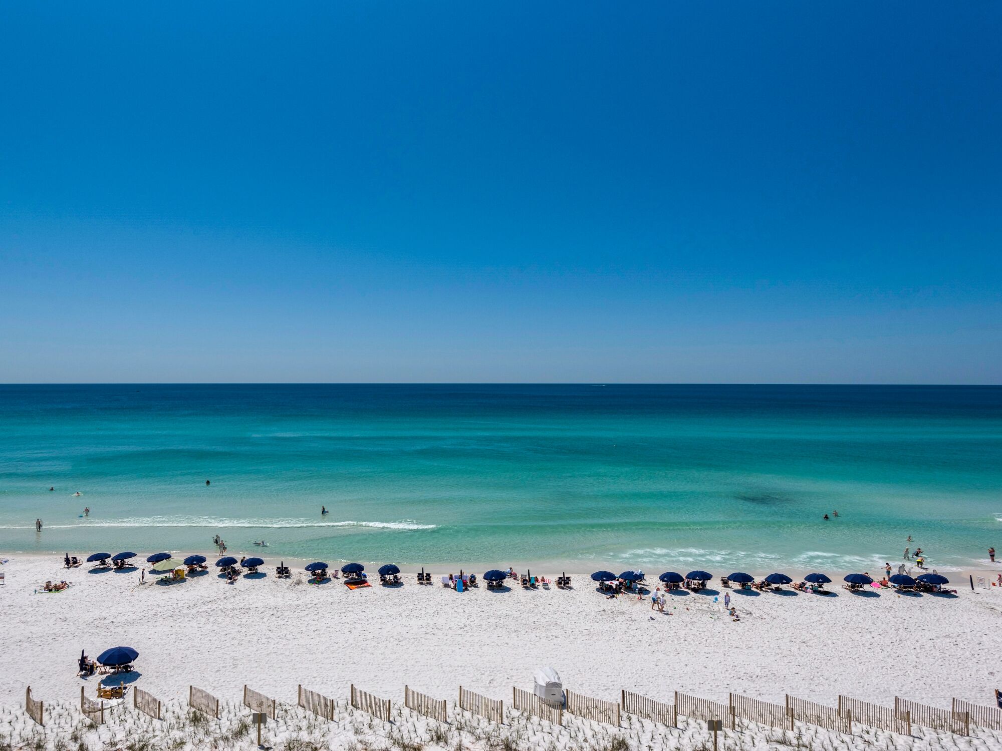 A beach with rows of umbrellas and beach chairs, a wooden fence, and the ocean in the background.